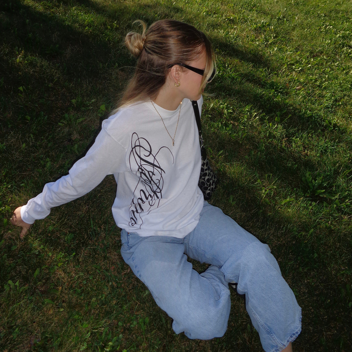 A woman sitting on a grassy lawn wearing a white long-sleeve t-shirt with a black graphic print on the back, light blue jeans, and black sunglasses.